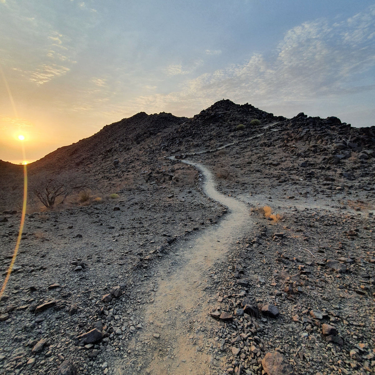 A dirt hiking trail winding up a rocky mountain slope during sunset, with a bright sun on the left and a blue sky with light clouds above.