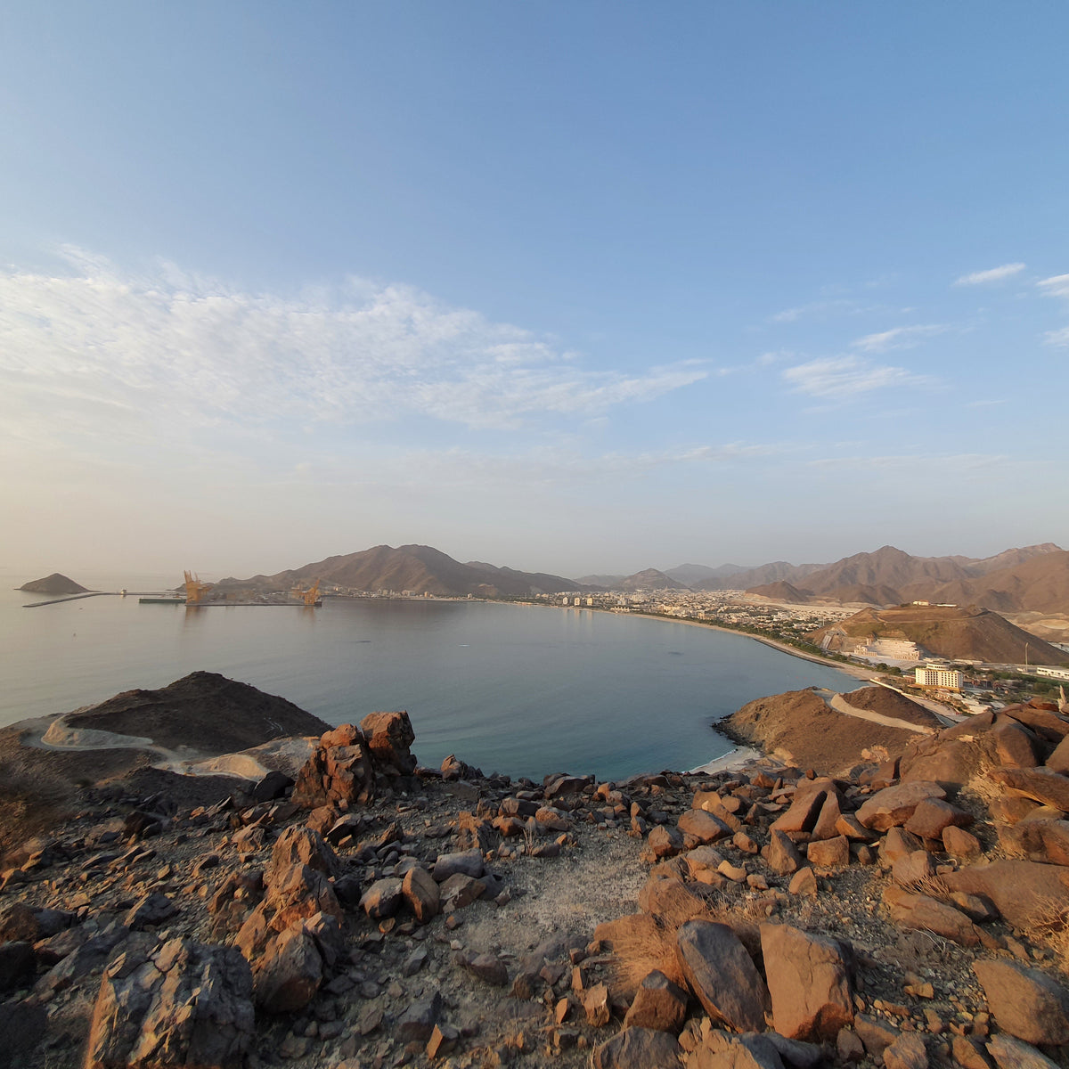 A scenic coastal landscape with rocky brown foreground, calm blue water, distant mountains, and a pale blue sky with light clouds.