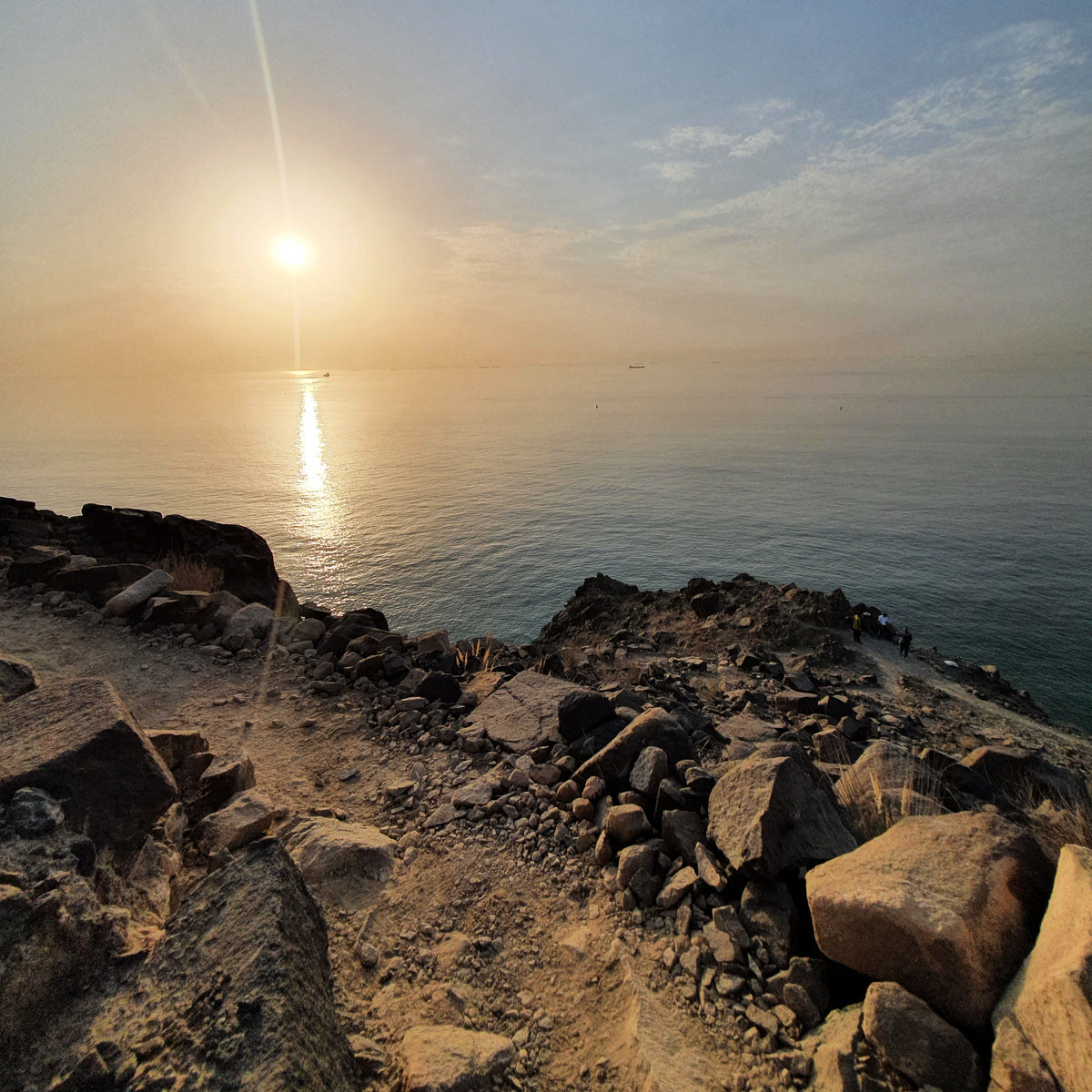 A scenic landscape photograph of a rocky shoreline at sunset, with the sun low over the horizon reflecting warm light on calm water under a blue sky.