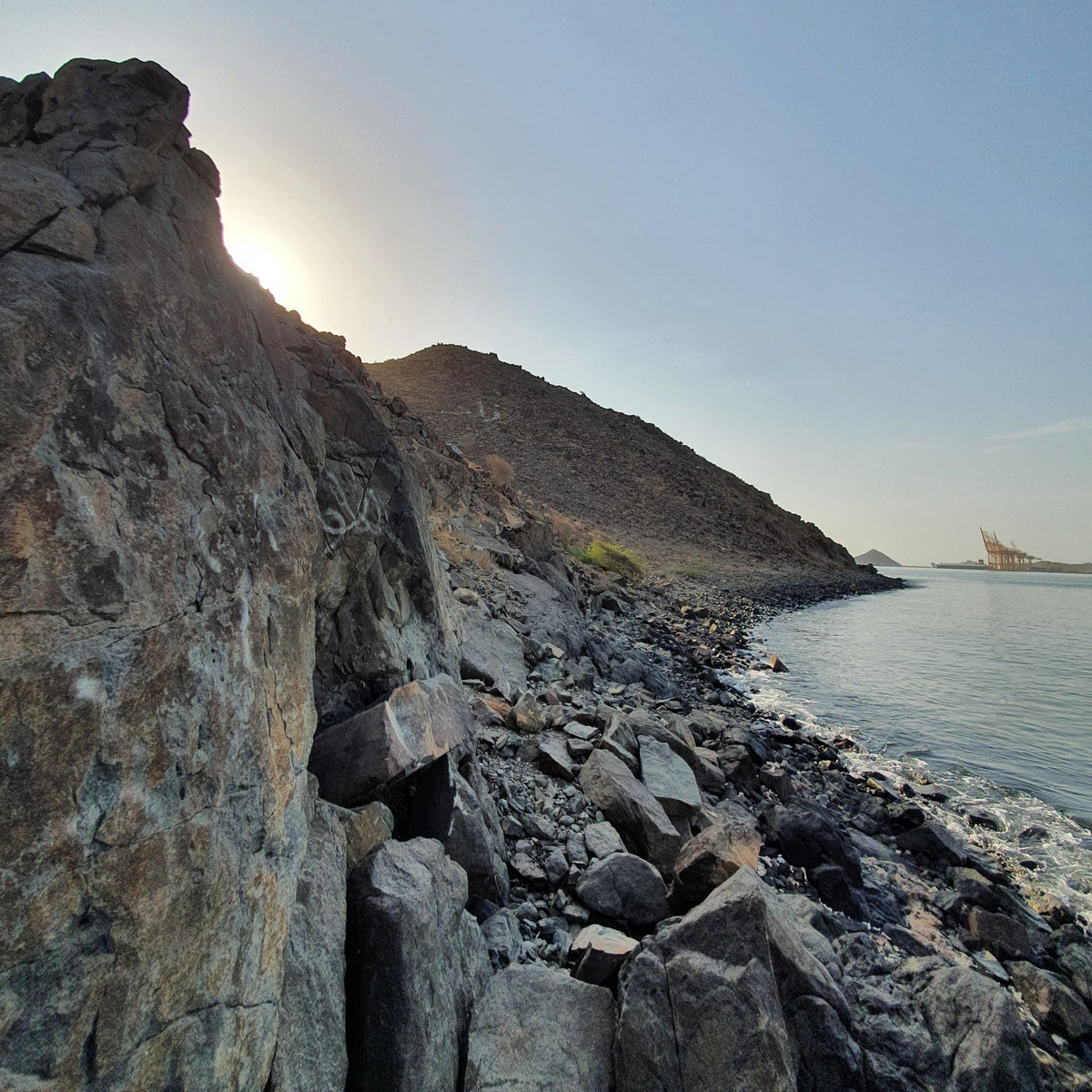 A scenic coastal landscape with dark rocky cliffs, a rocky shoreline, calm water, and a pale blue sky at sunrise or sunset.