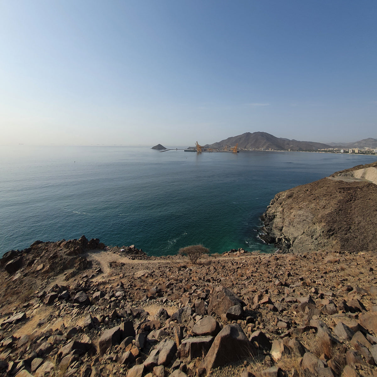 A scenic coastal landscape with a rocky brown cliff on the right, blue sea, and a distant island or peninsula under a clear blue sky.