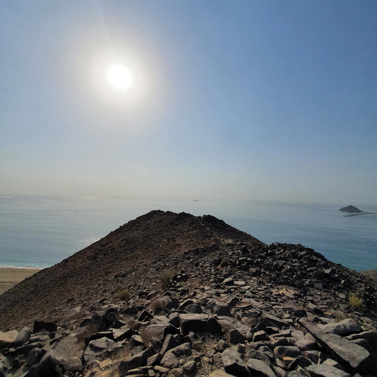 A rocky volcanic peak overlooking a calm blue sea under a bright sun and clear sky.