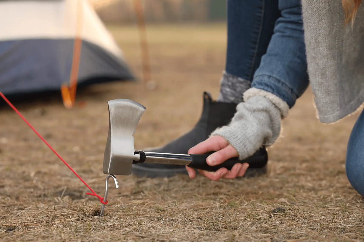 Person using Coghlan's Camp Axe to secure a tent stake in a camping setting