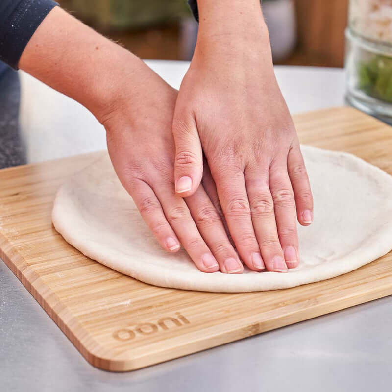 Hands pressing pizza dough on Ooni Pizza Prep Lid, a bamboo board.
