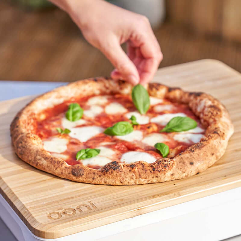 Person serving fresh wood-fired pizza directly on the Ooni Pizza Prep Lid