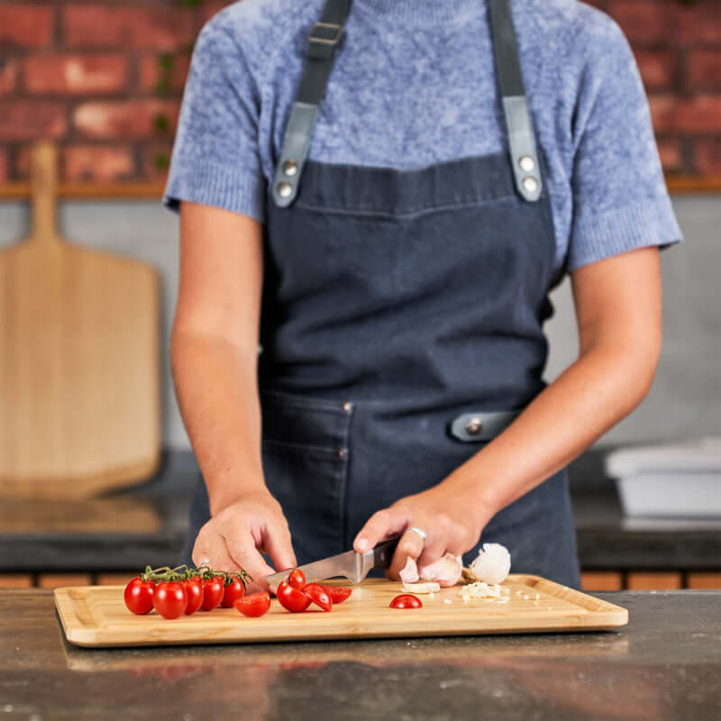Person preparing food on an Ooni Pizza Prep Lid - Bamboo Board with tomatoes and garlic in a kitchen setting.