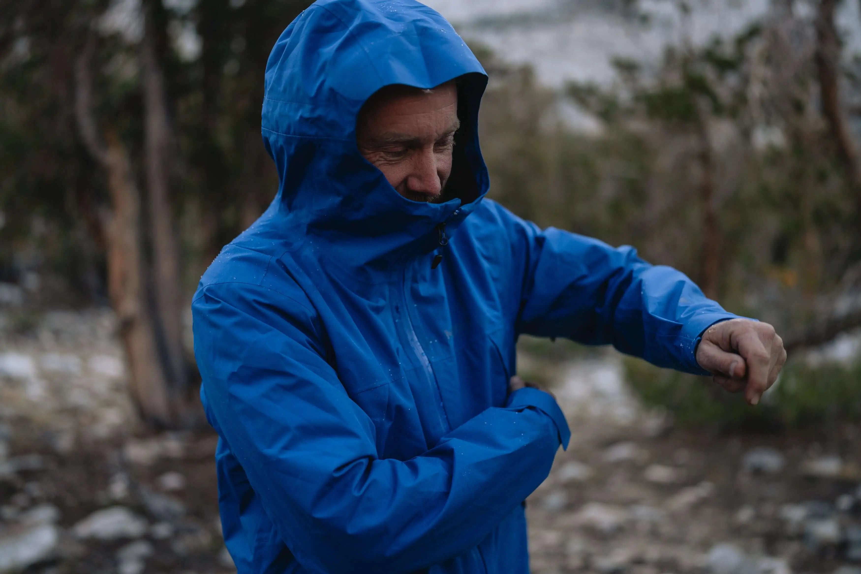 Man wearing a blue Marmot Gore-Tex Minimalist Pro Jacket in an outdoor setting, adjusting the sleeve during a hike.