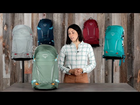 Load video: Woman showcasing various Osprey Hikelite 18 backpacks, highlighted in green, amidst a wooden backdrop. Ideal daypack for hiking.