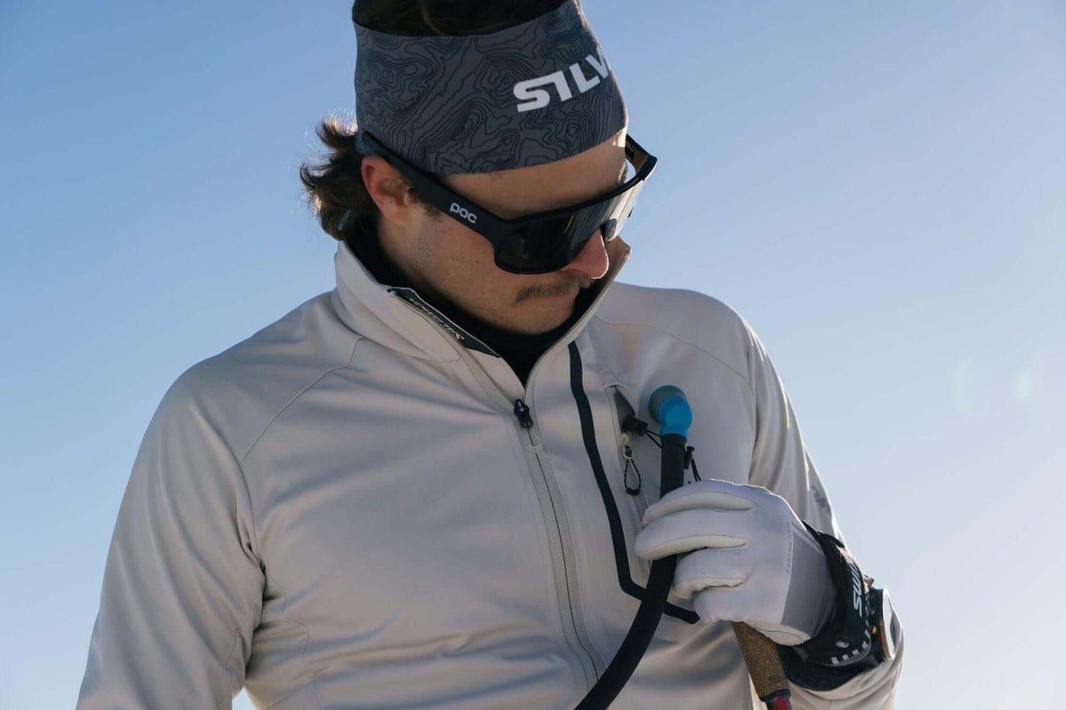 A close-up lifestyle shot of a skier wearing a light gray zip-up jacket, a gray patterned headband with 'STLV' in white, black sunglasses, and white gloves, holding a ski pole against a clear blue sky.