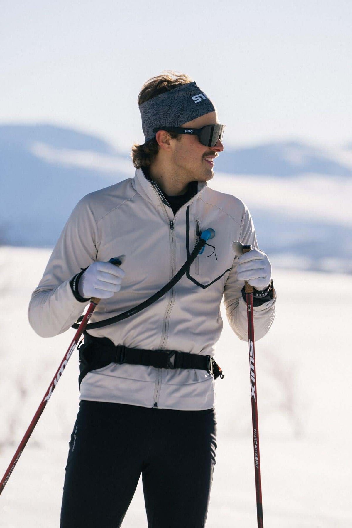 A skier wearing a white jacket, black pants, a gray headband, and sunglasses, holding ski poles in a snowy outdoor setting.
