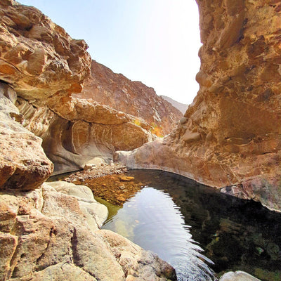 A scenic canyon landscape with towering tan and brown rock walls framing a narrow stream or pool of dark water under a pale blue sky.