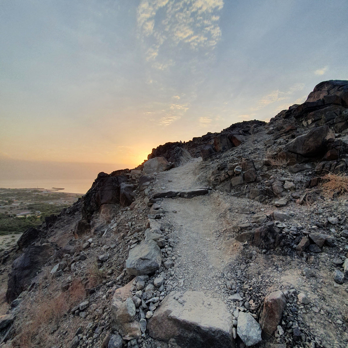 A rocky dirt trail winding up a steep hillside toward a sunset over the ocean, with scattered clouds in a blue sky.