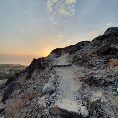 A rocky dirt trail winding up a steep hillside toward a sunset over the ocean, with scattered clouds in a blue sky.