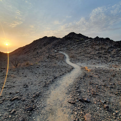 A dirt hiking trail winding up a rocky mountain slope during sunset, with a bright sun on the left and a blue sky with light clouds above.