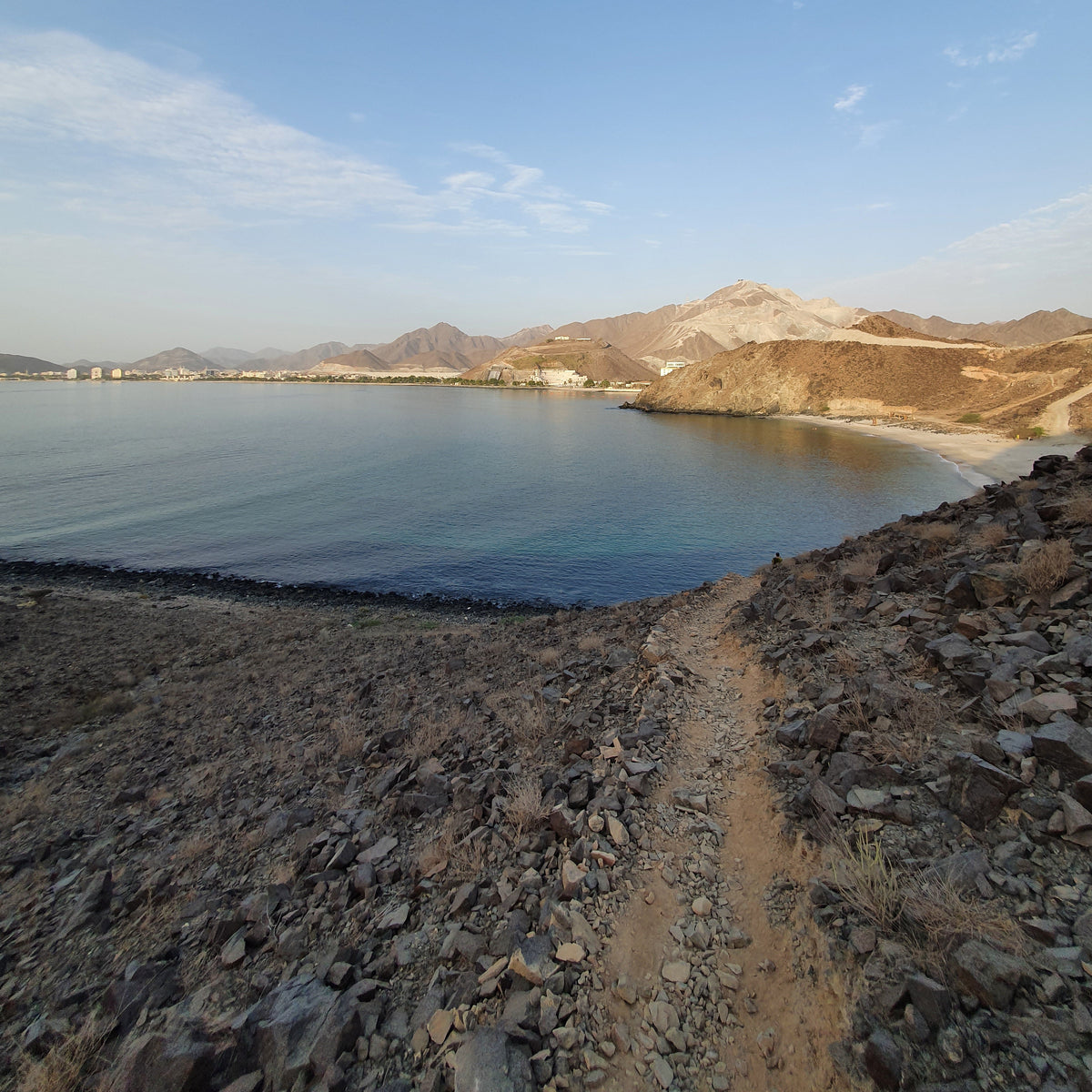 A scenic landscape photograph showing a rocky shoreline with a dirt path leading towards a calm blue body of water, with mountains in the background under a clear blue sky.