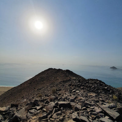 A rocky volcanic peak overlooking a calm blue sea under a bright sun and clear sky.