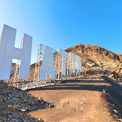 Large white metal letters spelling part of a destination sign mounted on a rocky desert hillside, with a dirt road in the foreground and blue sky above.