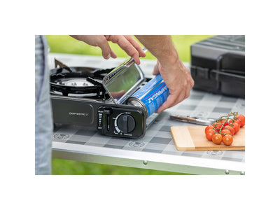 Person inserting Campingaz CP250 Isobutane Cartridge into portable stove on outdoor table.