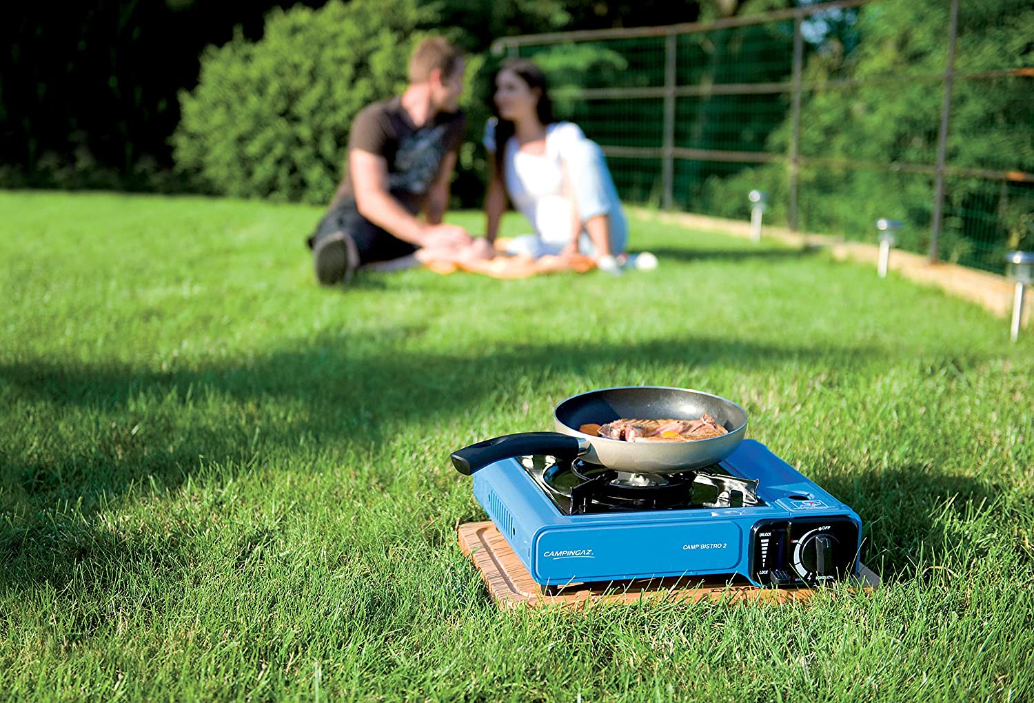 Campingaz Bistro 2 Portable Camp Stove on grass with food cooking, couple having picnic in background.