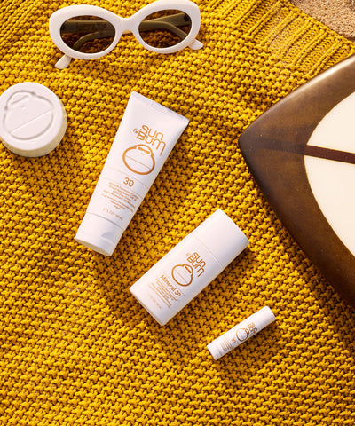Sun Bum Mineral SPF 30 Sunscreen Lip Balm with sunglasses on a yellow knit blanket by the beach.