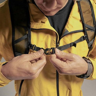 Close-up of a person wearing a yellow jacket and black backpack, adjusting a black strap buckle at the center chest.