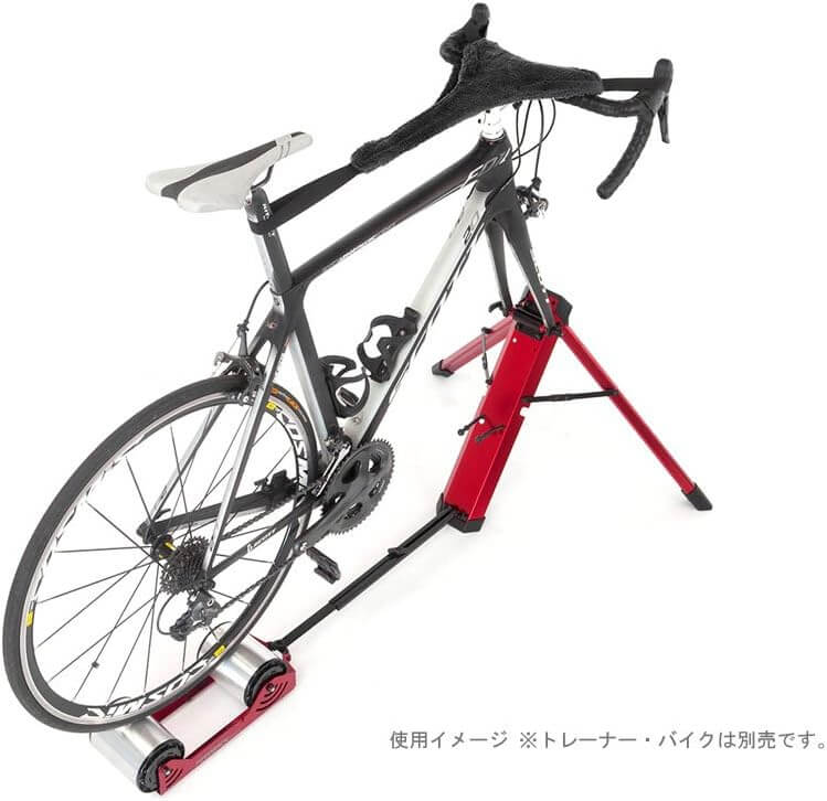 A road bicycle with a black handlebar tape and black saddle is mounted on a red and black indoor trainer stand against a white background. Japanese text appears at the bottom right.