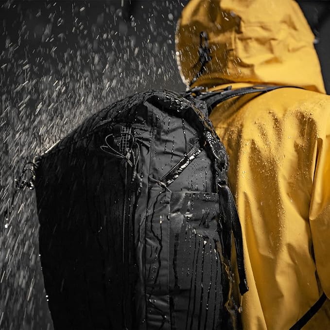 A black waterproof backpack shown from the back on a person wearing a yellow rain jacket, with heavy rain and water droplets spraying against a dark background.