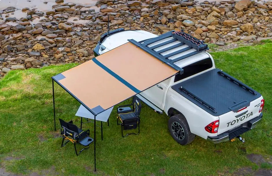 White truck equipped with Aluminium Awning with LED Light Kit - 2m x 2.5m - Black, set up in a campsite near a rocky shore.