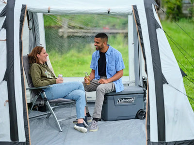 Two friends enjoying drinks inside a tent with a Coleman Convoy Wheeled Icebox Cooler - 65Qt / 61L beside them.