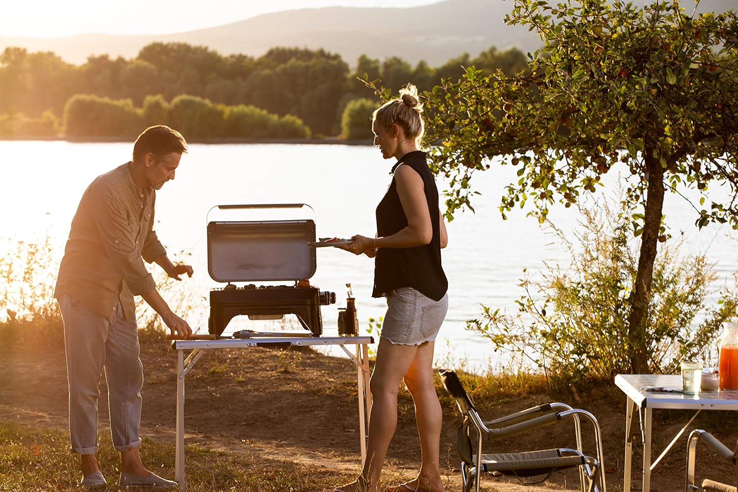 Couple grilling outdoors with Campingaz Attitude 2go CV Tabletop Gas Grill beside a lake at sunset.