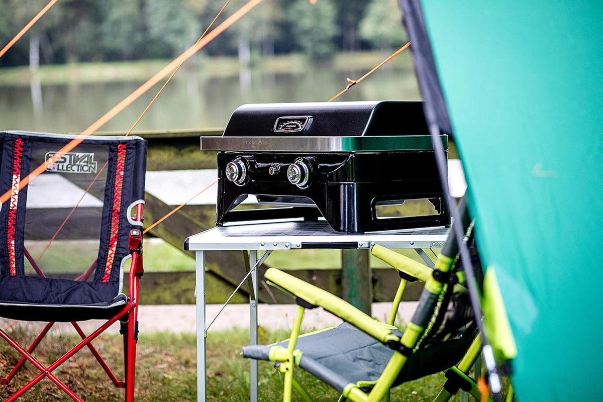 Campingaz Attitude 2100 LX Portable Gas Grill set up on a camping table by a lake, surrounded by camping chairs and a tent.