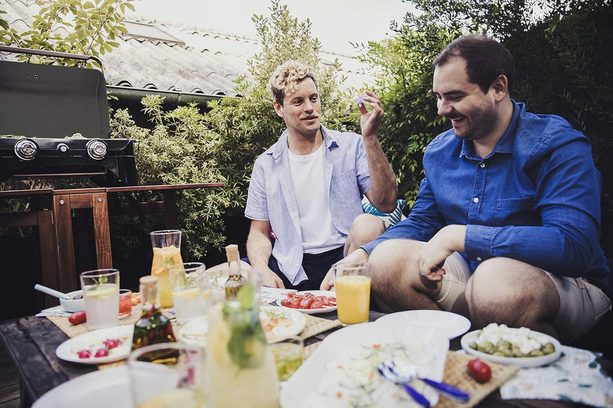 Two friends enjoying a meal outdoors near a Campingaz Attitude 2100 LX Portable Gas Grill on a sunny day.