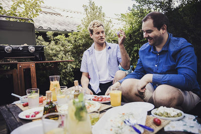 Two friends enjoying a meal outdoors near a Campingaz Attitude 2100 LX Portable Gas Grill on a sunny day.