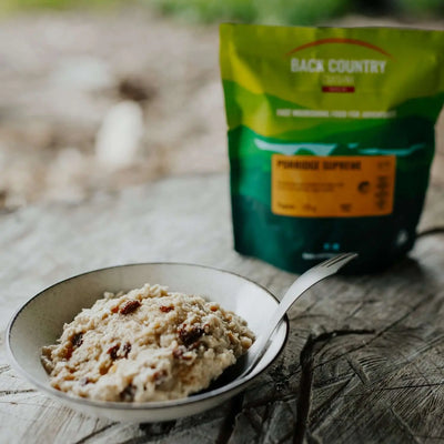 Back Country Cuisine Porridge Supreme camping breakfast with oats and dried fruit in a bowl, pouch in background on a wooden surface.