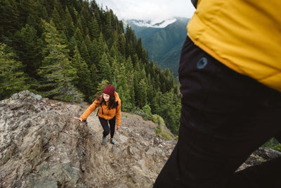 Woman hiking in Marmot Warmcube jacket on rocky trail surrounded by forest, showcasing outdoor performance and warmth.