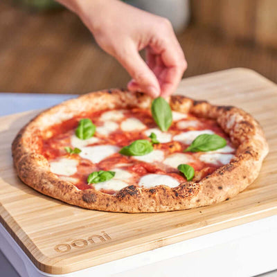 Person serving fresh wood-fired pizza directly on the Ooni Pizza Prep Lid