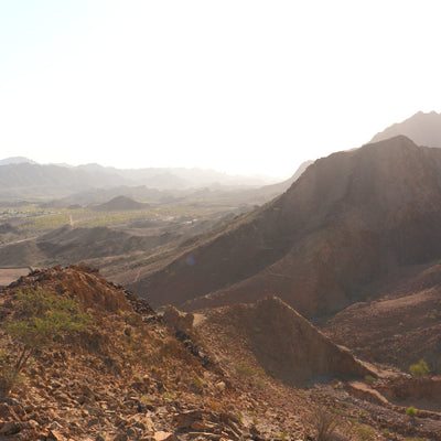 A scenic mountain landscape with rocky brown ridges, sparse vegetation, and distant hazy peaks under a bright sky.