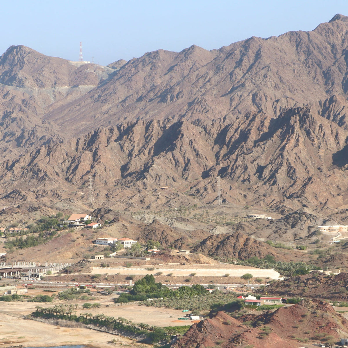 A scenic landscape photograph of rugged brown and tan mountains with a small cluster of buildings and greenery in the valley below under a clear blue sky.