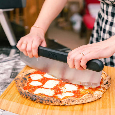 Person using Ooni Bamboo Pizza Peel and Serving Board to cut a freshly baked pizza with cheese topping in a kitchen setting.