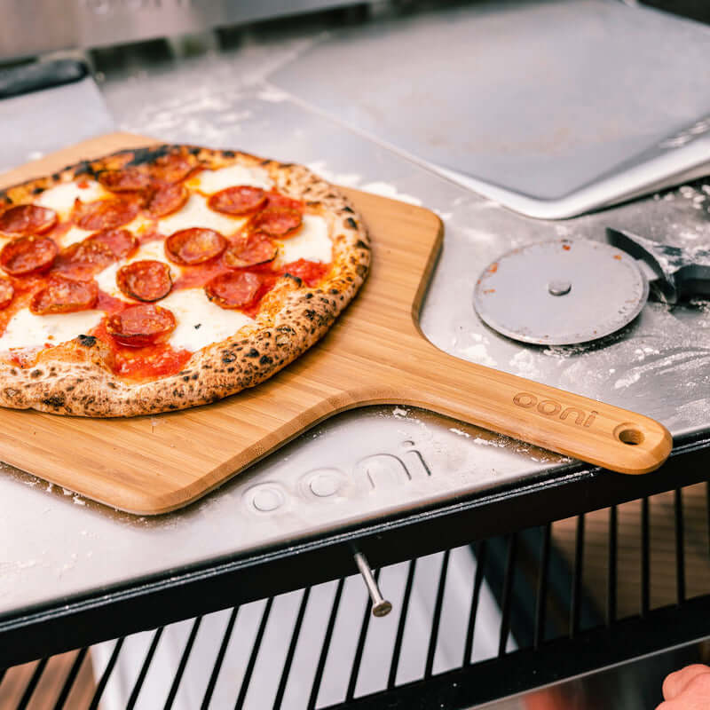 Ooni Bamboo Pizza Peel and Serving Board with pepperoni pizza next to a pizza cutter on a metal surface.