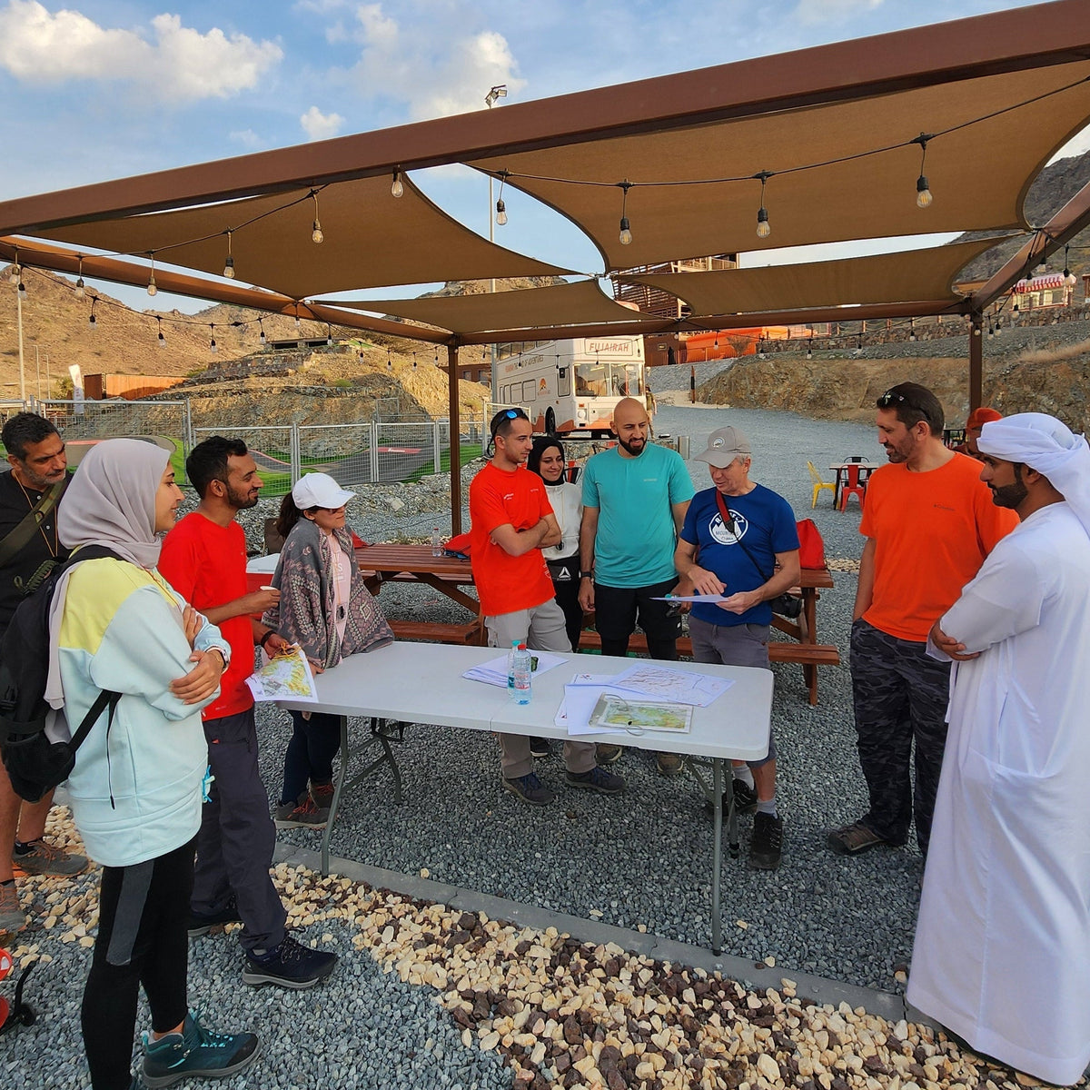 A group of people standing and talking outdoors under a large canopy structure. Several individuals are wearing traditional Middle Eastern clothing, including a white thobe and a white head covering, while others wear casual t-shirts and jeans.
