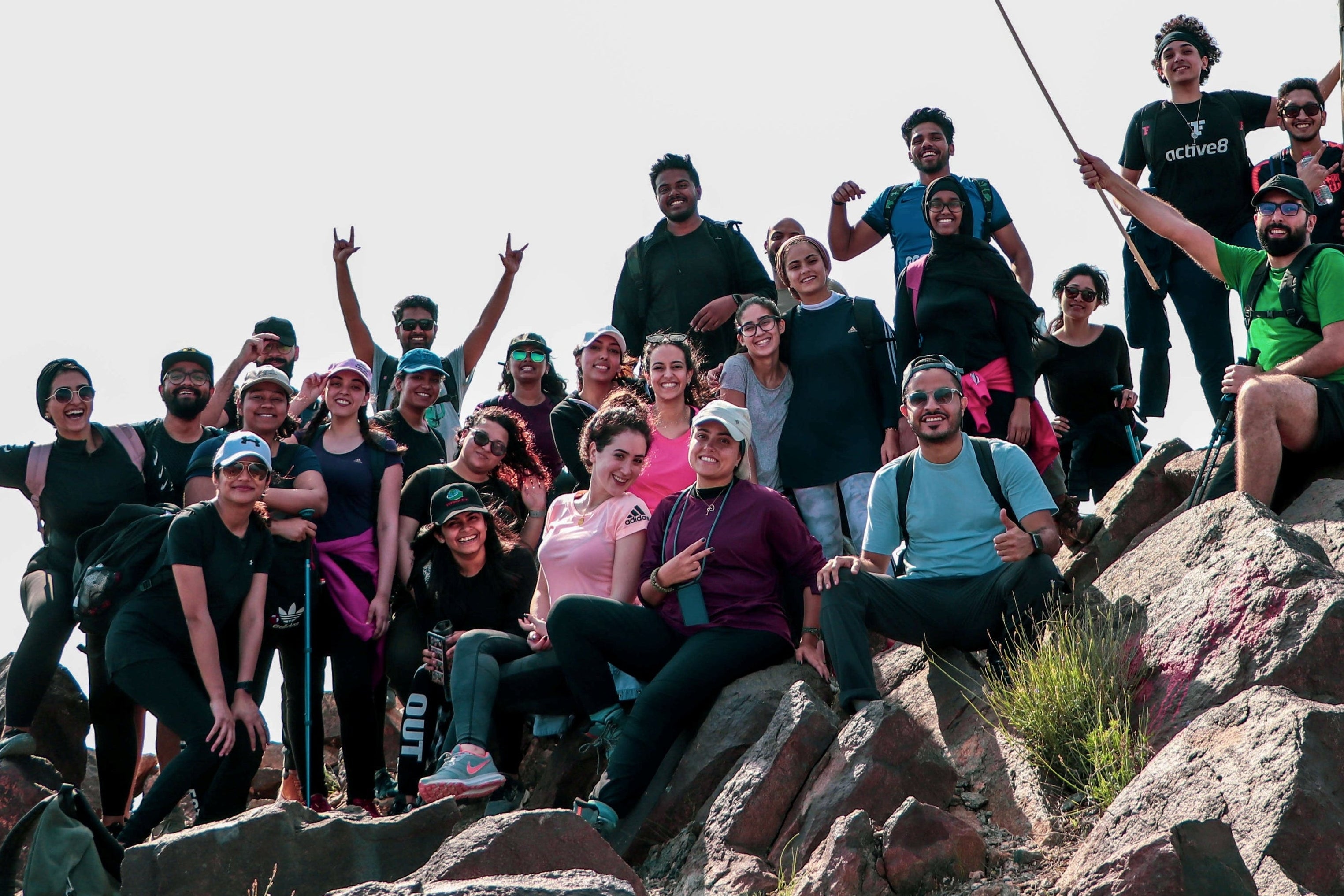 A large group of people posing together on rocky outdoor terrain under a bright sky, with two Palestinian flags being waved above them. This is a lifestyle shot featuring multiple people in a real environment.