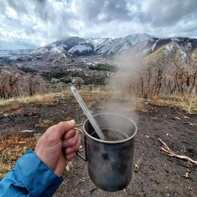 Person holding a steaming SilverAnt Ultralight Titanium Cup with Lid in a scenic mountainous background