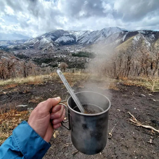 Person holding a steaming SilverAnt Ultralight Titanium Cup with Lid in a scenic mountainous background