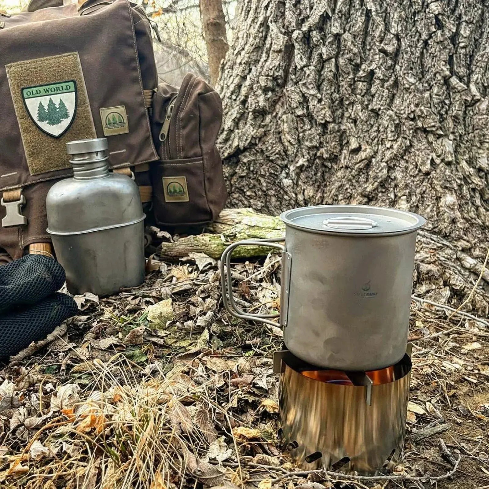 Camping gear featuring the SilverAnt Ultralight Titanium Stove Cross Stand and Windshield in a forest setting with backpack and canteen.
