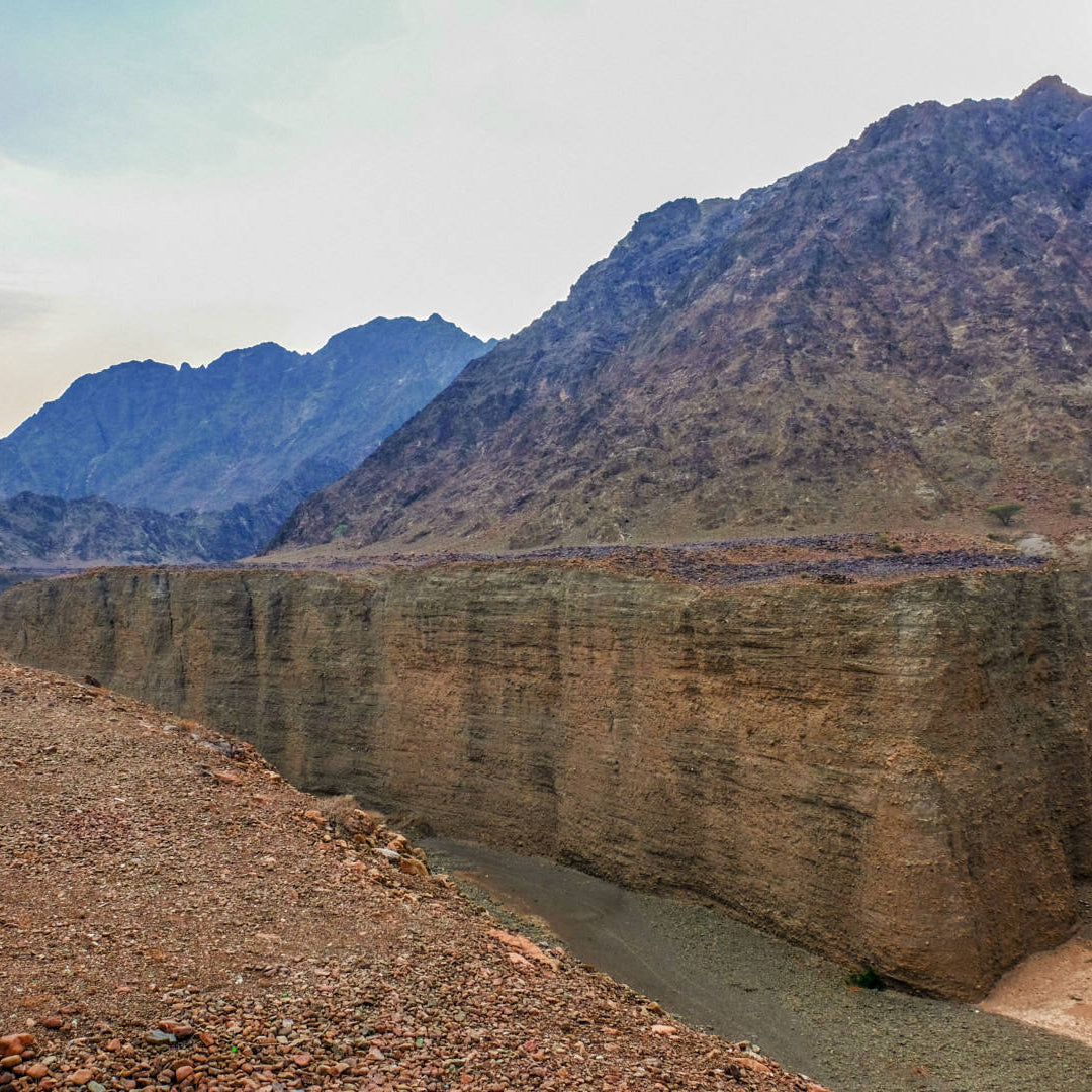 A dramatic natural canyon landscape with steep tan rock cliffs, a narrow river below, and rugged mountains under a pale sky.