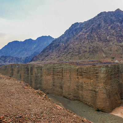 A dramatic natural canyon landscape with steep tan rock cliffs, a narrow river below, and rugged mountains under a pale sky.