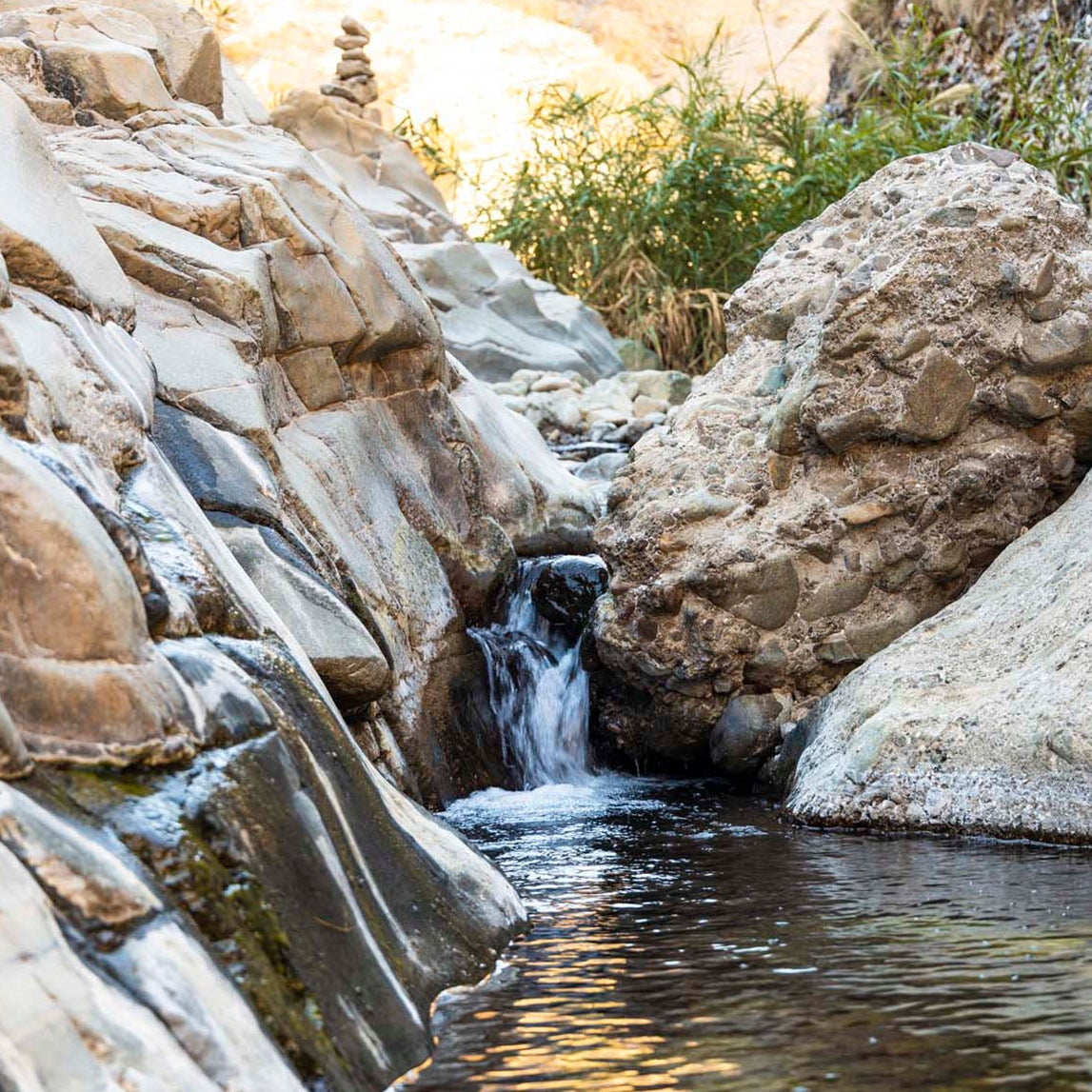 A natural rocky stream with a small waterfall flowing into a pool, surrounded by tan and gray boulders with some green plants in the background.