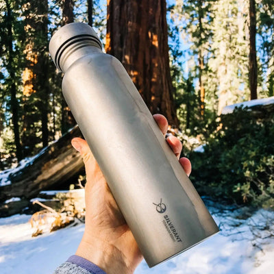 Hand holding SilverAnt Round Titanium Water Bottle - 27 oz in forest backdrop, durable and lightweight for outdoor adventures.
